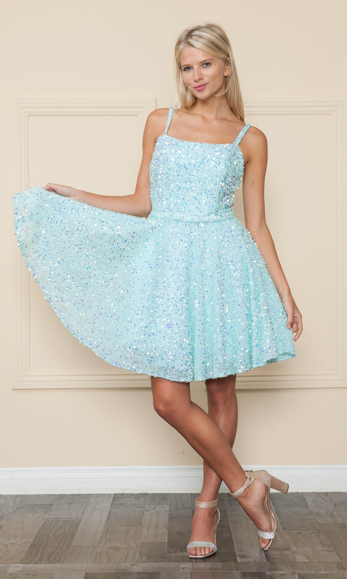 A woman wearing the Short A-Line Sequin Homecoming Dress 8930 in light blue poses in open-toe heels, holding out the skirt with one hand against a beige wall.